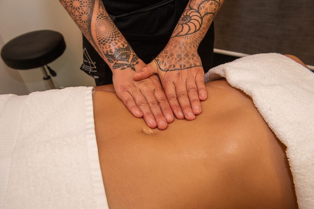 Hands of a tattooed therapist performing gentle abdominal lymphatic drainage massage on a client lying under a white towel.